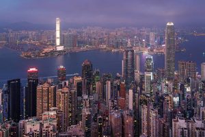 A panoramic view of Hong Kong captured from The Peak, showcasing the iconic skyline of Central with its dense cluster of modern skyscrapers and Victoria Harbour stretching toward Kowloon in the distance.